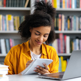 Mujer escribiendo en un bloc de notas mientras está sentada frente a una computadora portátil